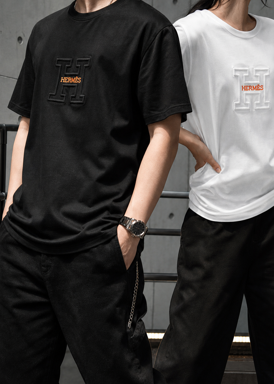 Two models wearing oversized graphic t-shirts with a large 'H' logo and 'HERNICS' text. One model wears a black t-shirt and black pants, while the other wears a white t-shirt and black pants.