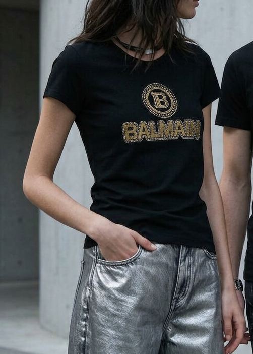 A man and a woman wearing matching black Balmain t-shirts with a gold logo, standing in front of a concrete wall.