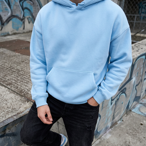 A person wearing a light blue pullover hoodie with a front kangaroo pocket, paired with black jeans and white sneakers, standing outdoors against a graffiti-covered wall.